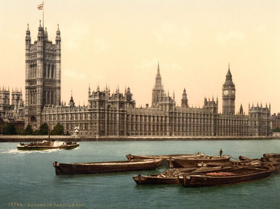 Hand-coloured view of the Gothic Houses of Parliament and the clock tower of Big Ben across the River Thames, with barges moored in the foreground