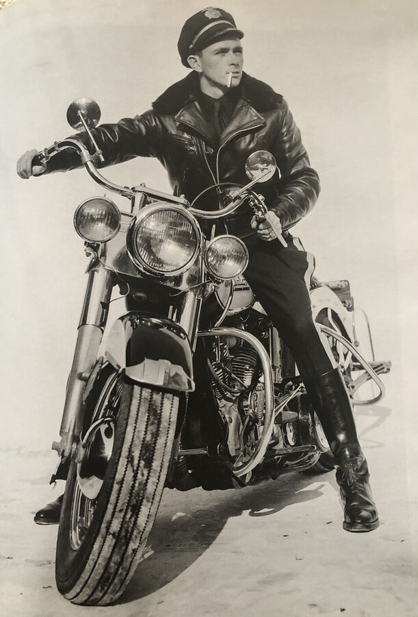 Black and white photograph of a man in a leather jacket, peaked cap and boots sitting on a large motorcycle with a cigarette