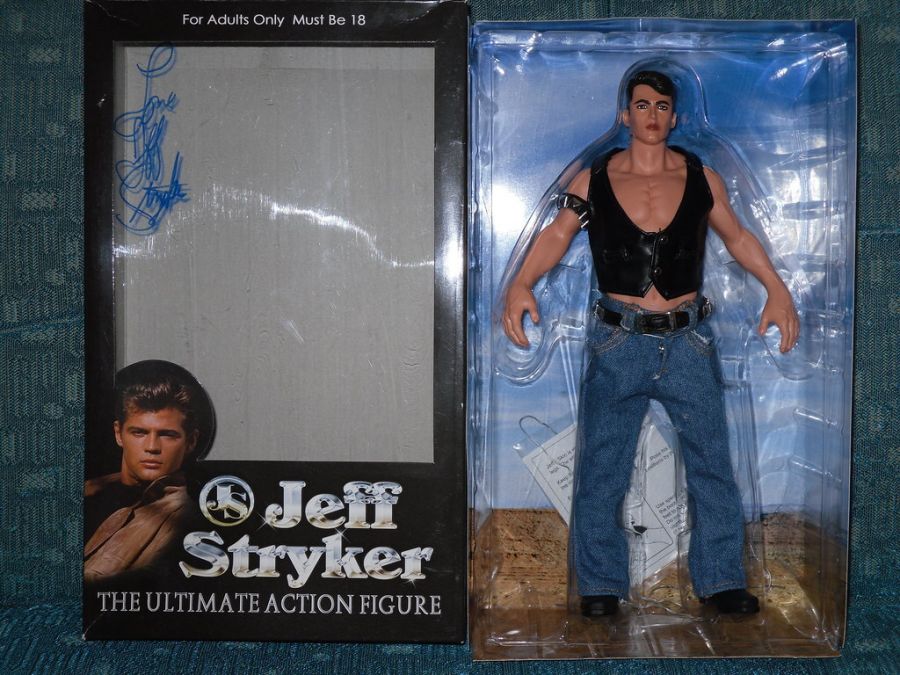 A Jeff Stryker action figure in a leather vest and jeans inside its open box, which carries his signature and a printed portrait of him