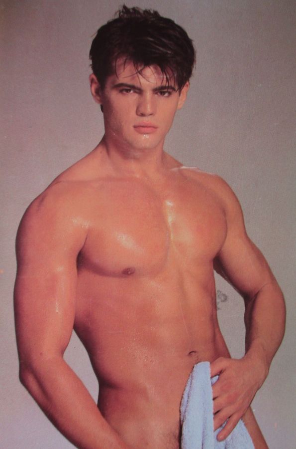 A young dark-haired man with wet, swept-down hair and a muscular, oiled chest stands against a grey backdrop, looking straight at the camera and holding a pale blue towel in front of his hips