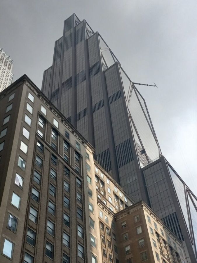 A tall dark glass skyscraper with angled steel supports rising behind an older brown brick building under a grey sky