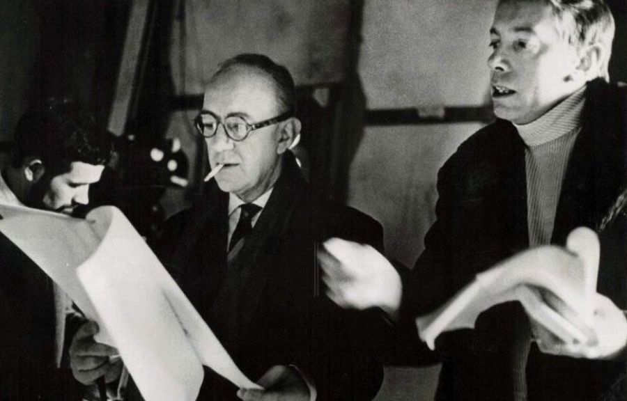 Black and white photograph of three men studying the pages of a script, an older man in glasses with a cigarette in the centre and a man in a dark coat and roll-neck sweater on the right