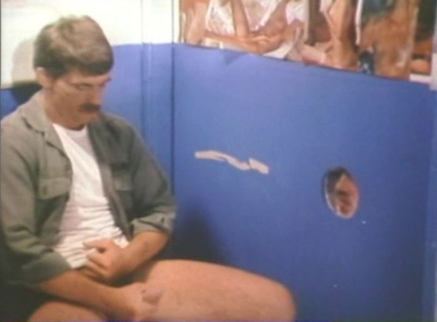 A moustached man in glasses and a work shirt sits with his trousers down in a booth painted blue, beside a glory hole in the partition