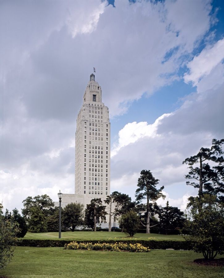 A tall Art Deco skyscraper of pale stone rising above lawns and pine trees under a sky with white clouds