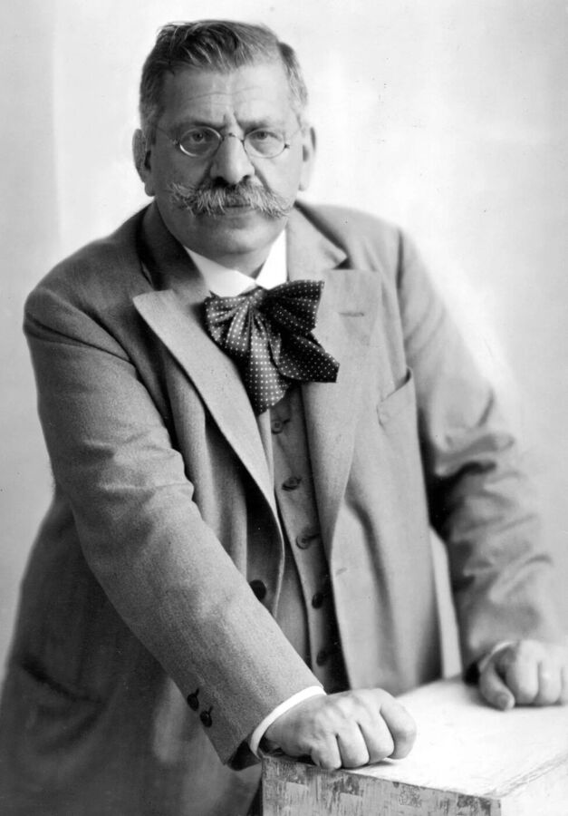 Black and white photograph of a heavy-set man with a thick moustache, round glasses and a large bow tie, leaning on a table