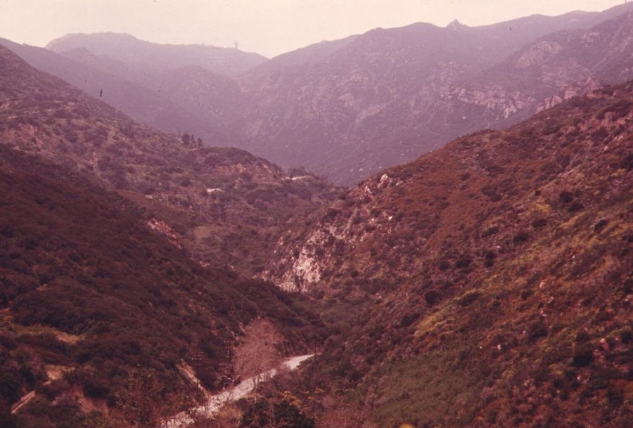 A deep green canyon winding between steep scrub-covered mountains under a hazy pink sky