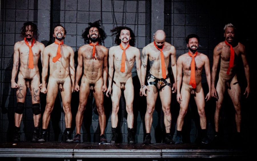 Seven naked actors in a row on a dark stage, wearing only orange ties and boots, grimacing at the audience with their mouths held open