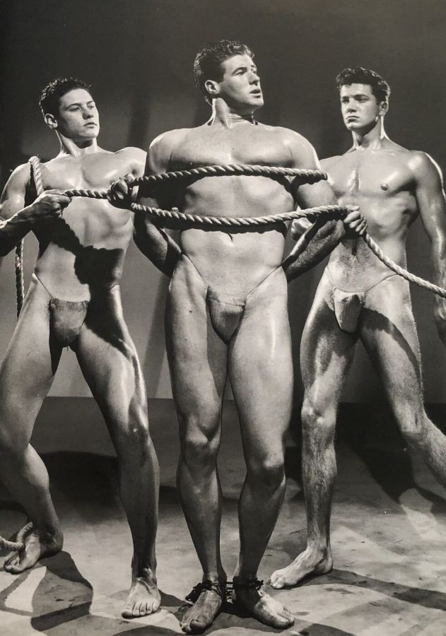 Black and white studio photograph of three muscular men in posing straps, the man in the centre wrapped in a thick rope held by the two others
