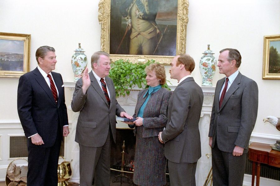 Five people in suits in the Oval Office: a man raises his right hand to take an oath while a woman holds a Bible, watched by President Reagan and Vice President Bush