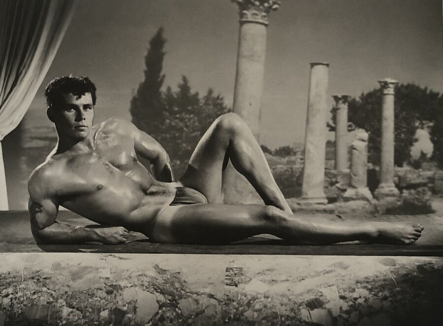 Black and white photograph of a muscular man in a posing strap reclining on a ledge in front of a painted backdrop of classical ruins and columns