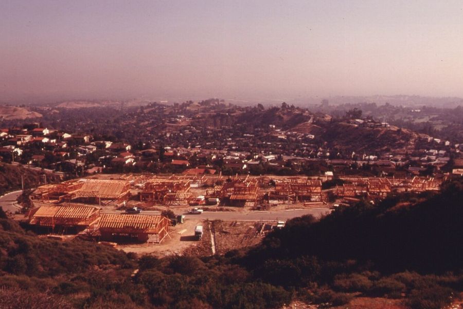 View from a hilltop of rows of new houses under construction in a cleared valley, with wooded hills and a hazy sky beyond