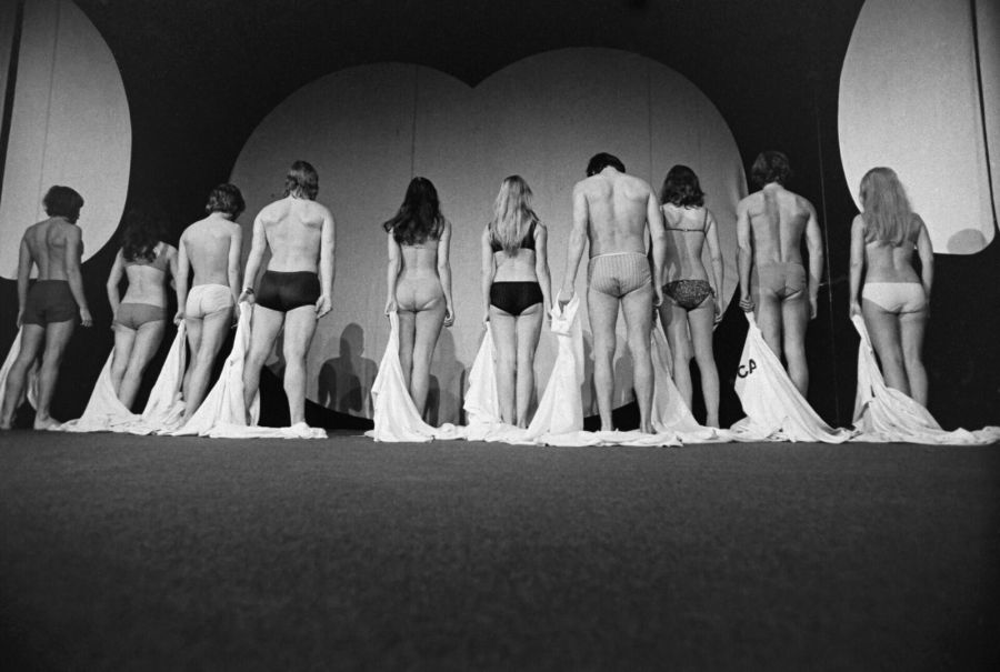 Black and white photograph of a row of men and women seen from behind on a stage, in underwear, holding the white robes they have just taken off
