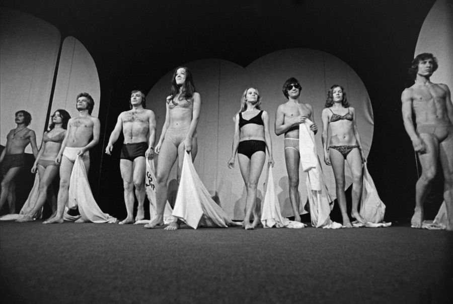 Black and white photograph of the same cast facing the audience in underwear and bikinis, the white robes at their feet