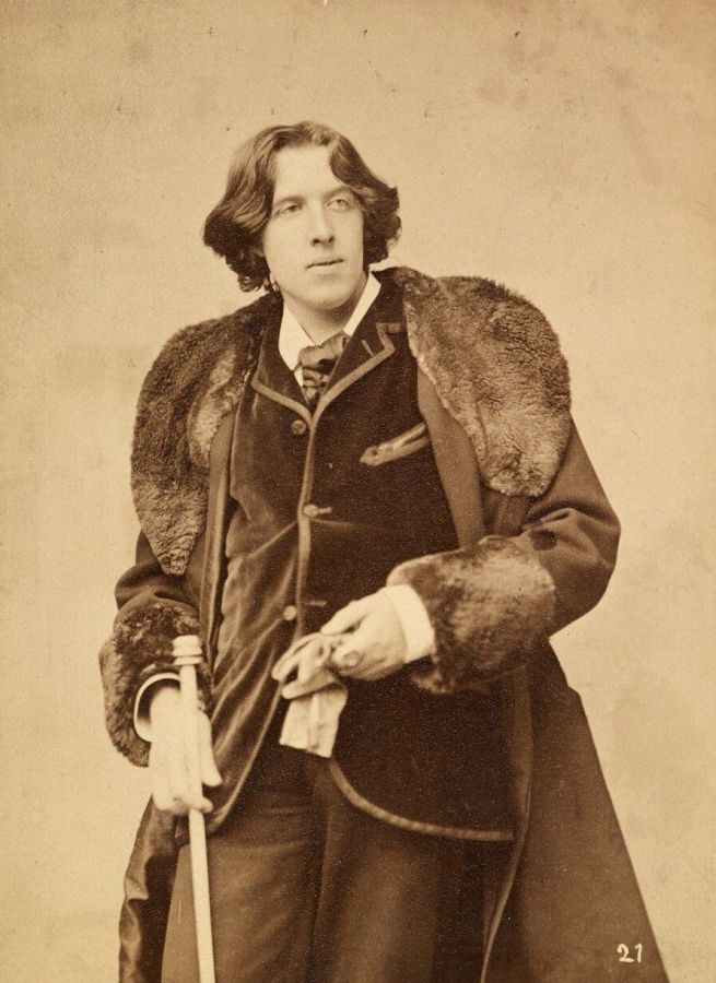 Full-length photograph of Oscar Wilde with long hair, standing in a fur-trimmed overcoat, holding a cane and a pair of gloves