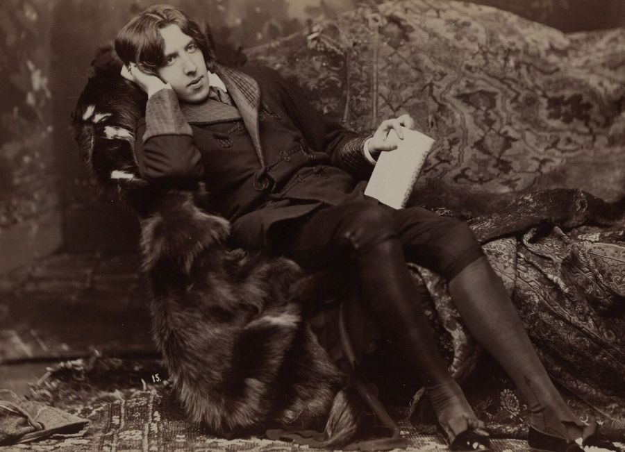 Sepia photograph of Oscar Wilde in knee breeches reclining on a couch draped with furs, holding a book