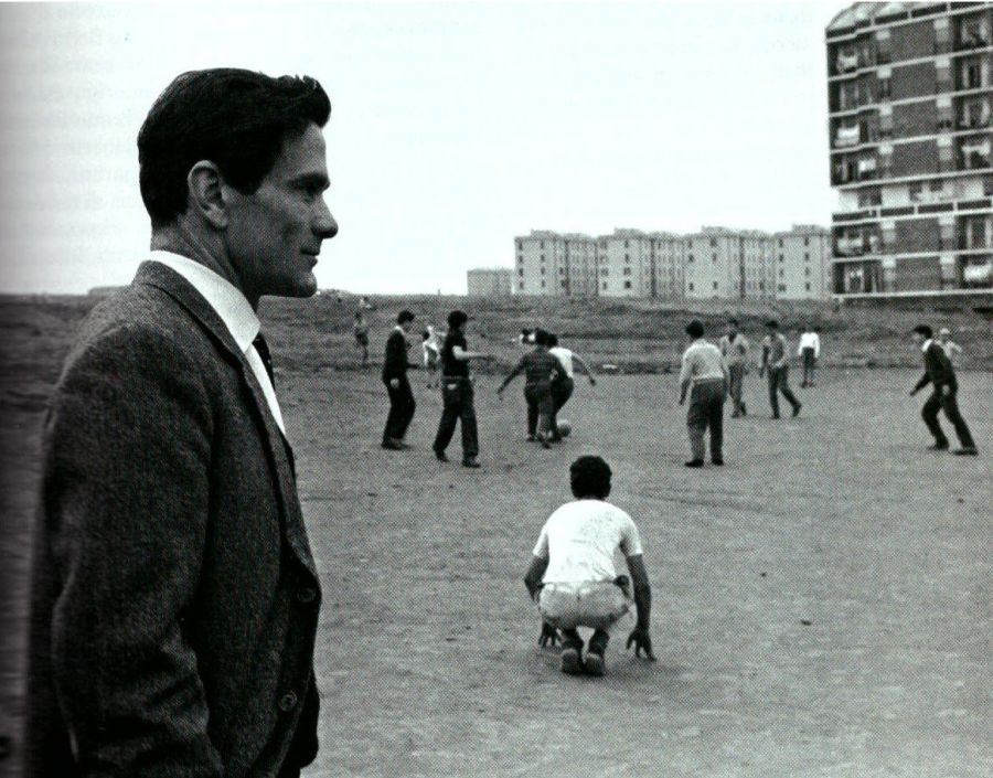 A dark-haired man in a tweed jacket and tie stands in profile in the foreground of a dusty open field where boys play football, with blocks of flats behind them