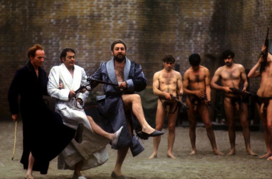 Three middle-aged men in dressing gowns dance with high kicks in a walled courtyard while four young men wearing only loincloths and holding rifles stand watching