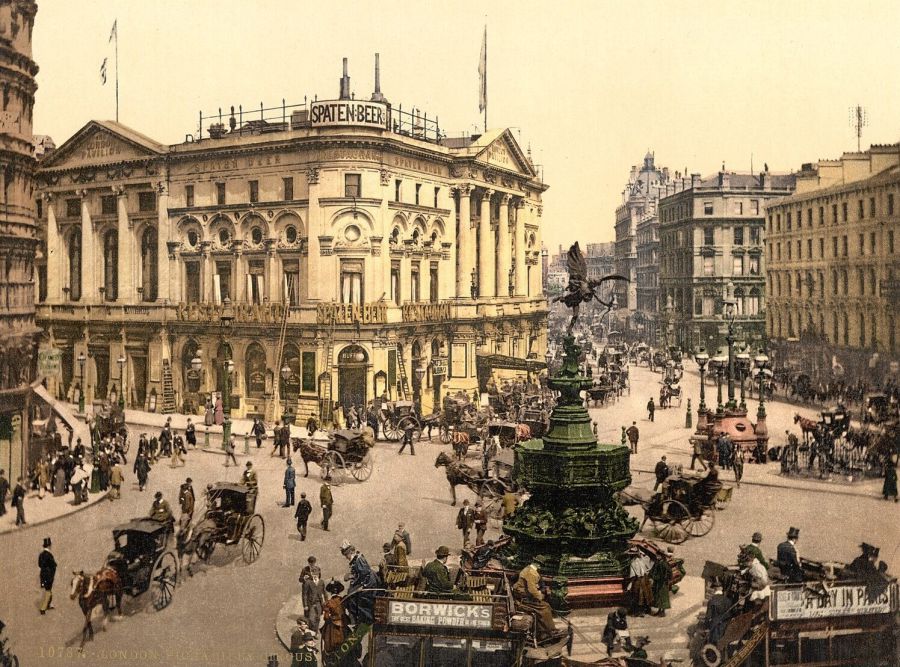 A busy London square in colour, crowded with horse-drawn omnibuses and cabs around a fountain, with tall stone buildings behind