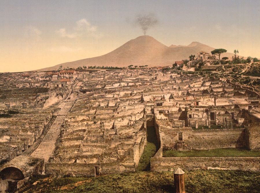 Colour print of the excavated streets and roofless houses of Pompeii with Vesuvius smoking in the background
