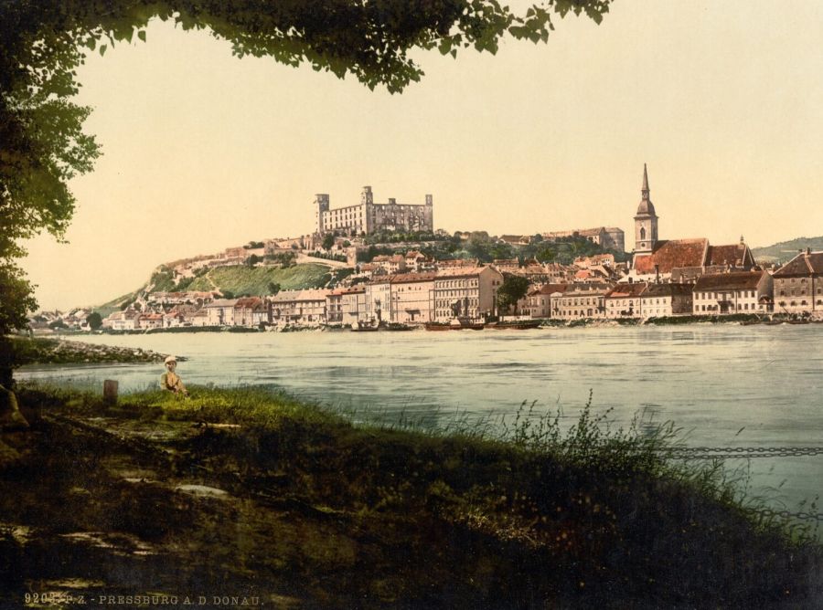 A colour view across the Danube of Bratislava, with the square castle on its hill and the spire of the cathedral above the houses on the riverbank