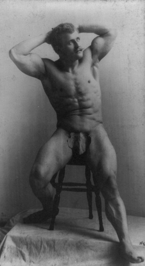 Photograph of Eugen Sandow sitting on a stool with his hands behind his head, naked except for a fig leaf, his legs apart and muscles tensed