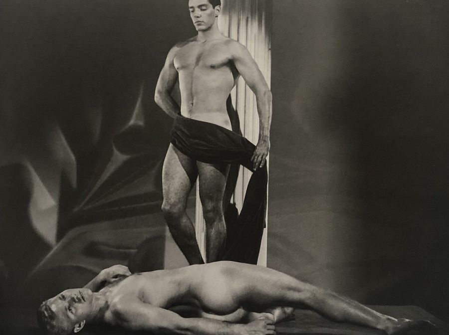 Black and white studio photograph of a muscular man standing and holding a dark drape in front of his groin while a second muscular man lies naked on the floor at his feet