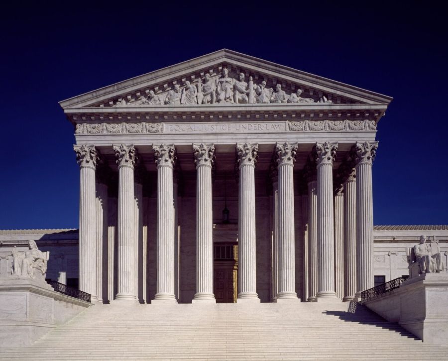 The white marble front of the Supreme Court building, with a row of tall Corinthian columns, a carved pediment and wide steps, against a deep blue sky
