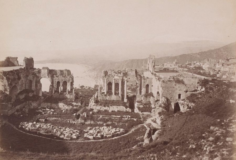 Old sepia photograph of the ruined ancient theatre of Taormina, with the bay and the town on the hillside behind it