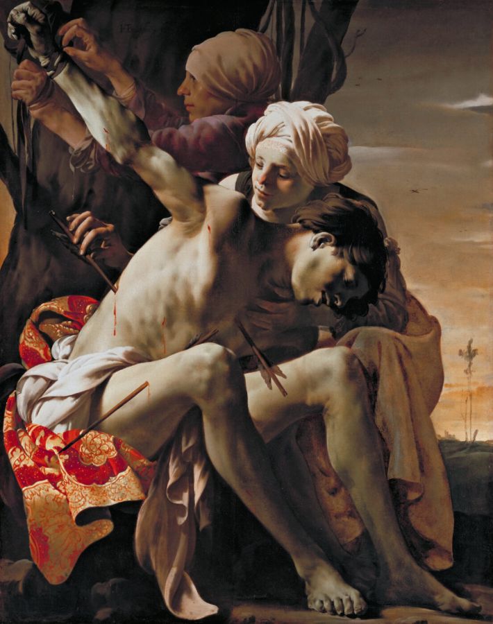 Two women untie a nearly naked, unconscious man from a tree and pull out an arrow, his arm still bound above him, a red cloth beneath him