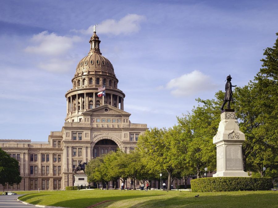 The domed pink granite Texas State Capitol behind green lawns and trees, with a bronze statue on a tall pedestal to the right, under a blue sky with clouds