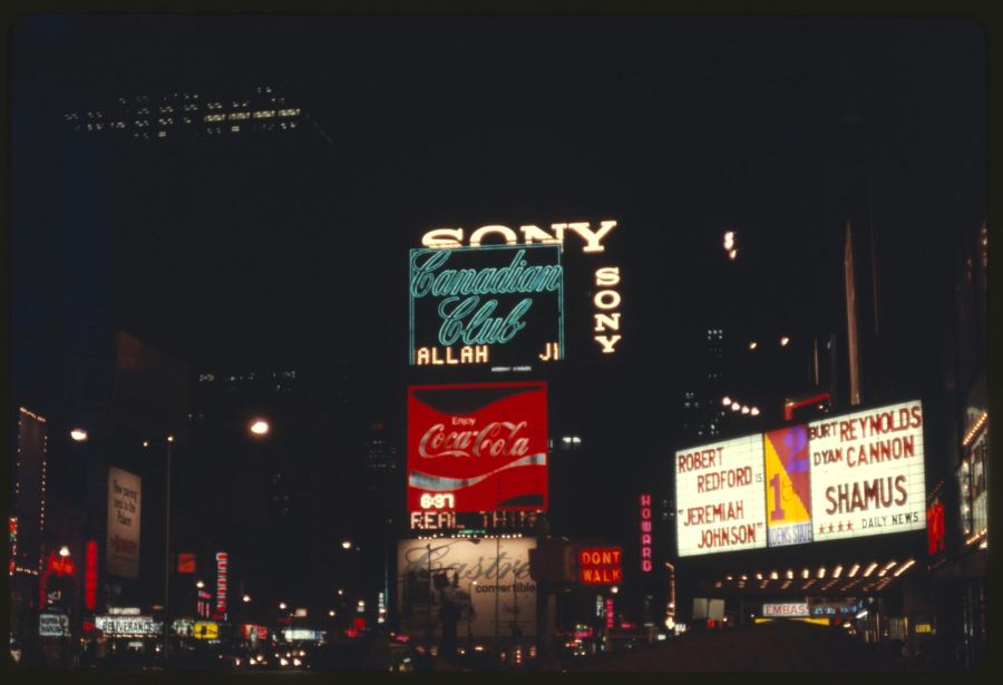 Times Square at night with neon signs for Sony, Canadian Club and Coca-Cola, and a lit cinema marquee announcing Burt Reynolds and Dyan Cannon in Shamus next to Robert Redford in Jeremiah Johnson