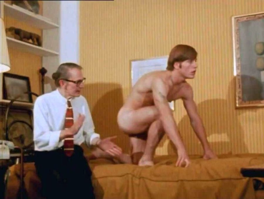 Film still of Joe Dallesandro naked, crouching on a sofa bed in a yellow room, while an elderly man in a shirt and tie looks at him