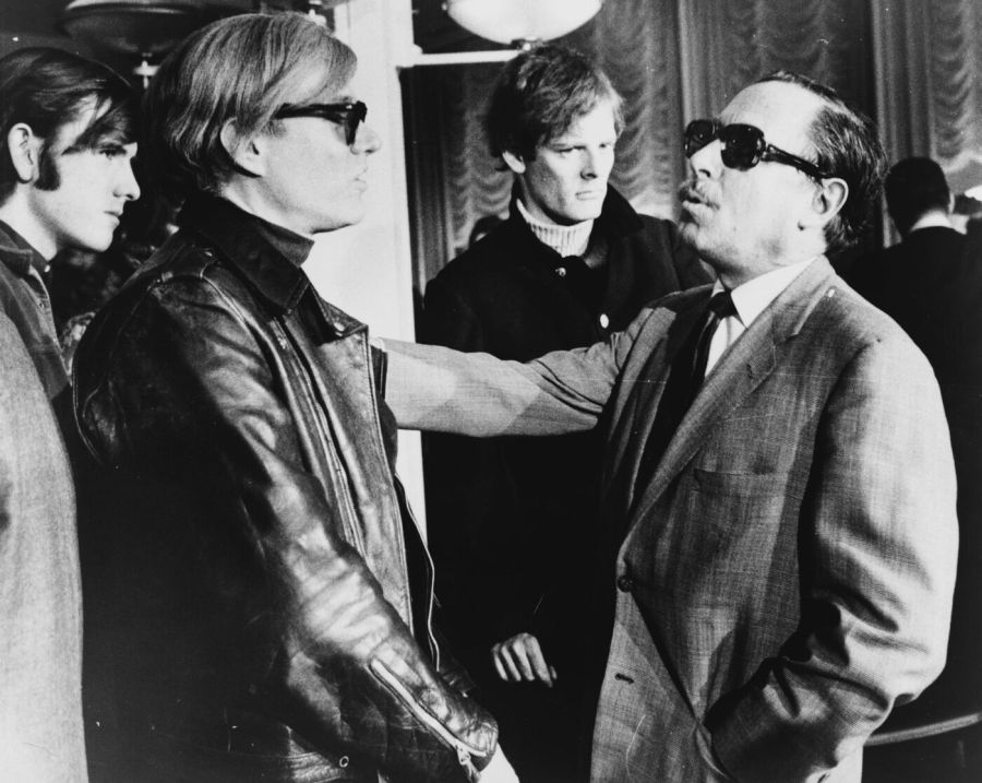 Andy Warhol in a leather jacket and dark glasses talking with Tennessee Williams in a suit and dark glasses, with Paul Morrissey standing behind them and another young man at the left