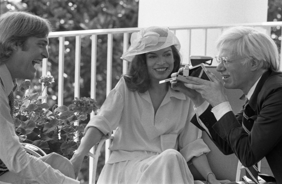 Andy Warhol, with pale hair and glasses, holding a Polaroid camera up to a woman in a wide-brimmed hat sitting beside a young man on a white balcony with plants