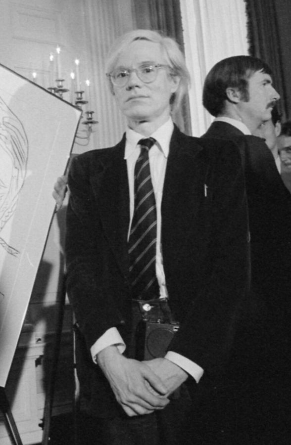Andy Warhol, with pale hair and glasses, in a dark suit and striped tie, standing with his hands clasped at a reception in a White House room