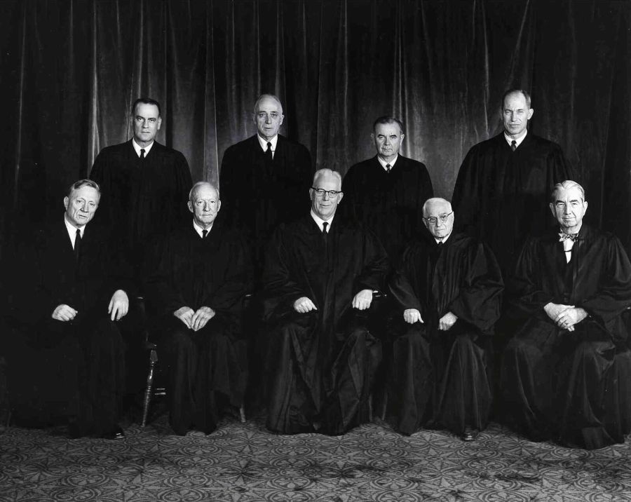 Group portrait of nine judges in black robes, five seated in front and four standing behind them, against a dark curtain