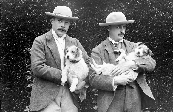 Two men in suits and straw hats standing in a garden, each holding a small dog in his arms